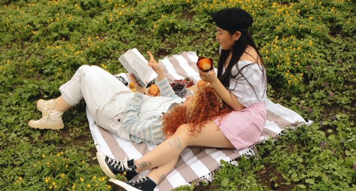 an Asian and white woman sitting and reading a book on a picnic blanket