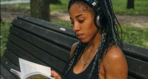 brown-skinned Black woman with braids reading with headphones on while sitting on a park bench