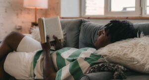 brown-skinned young man with locs is lying down and reading a book on the couch