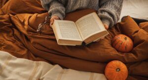fair-skinned hands holding a book open next to a blanket, glasses, two pumpkins