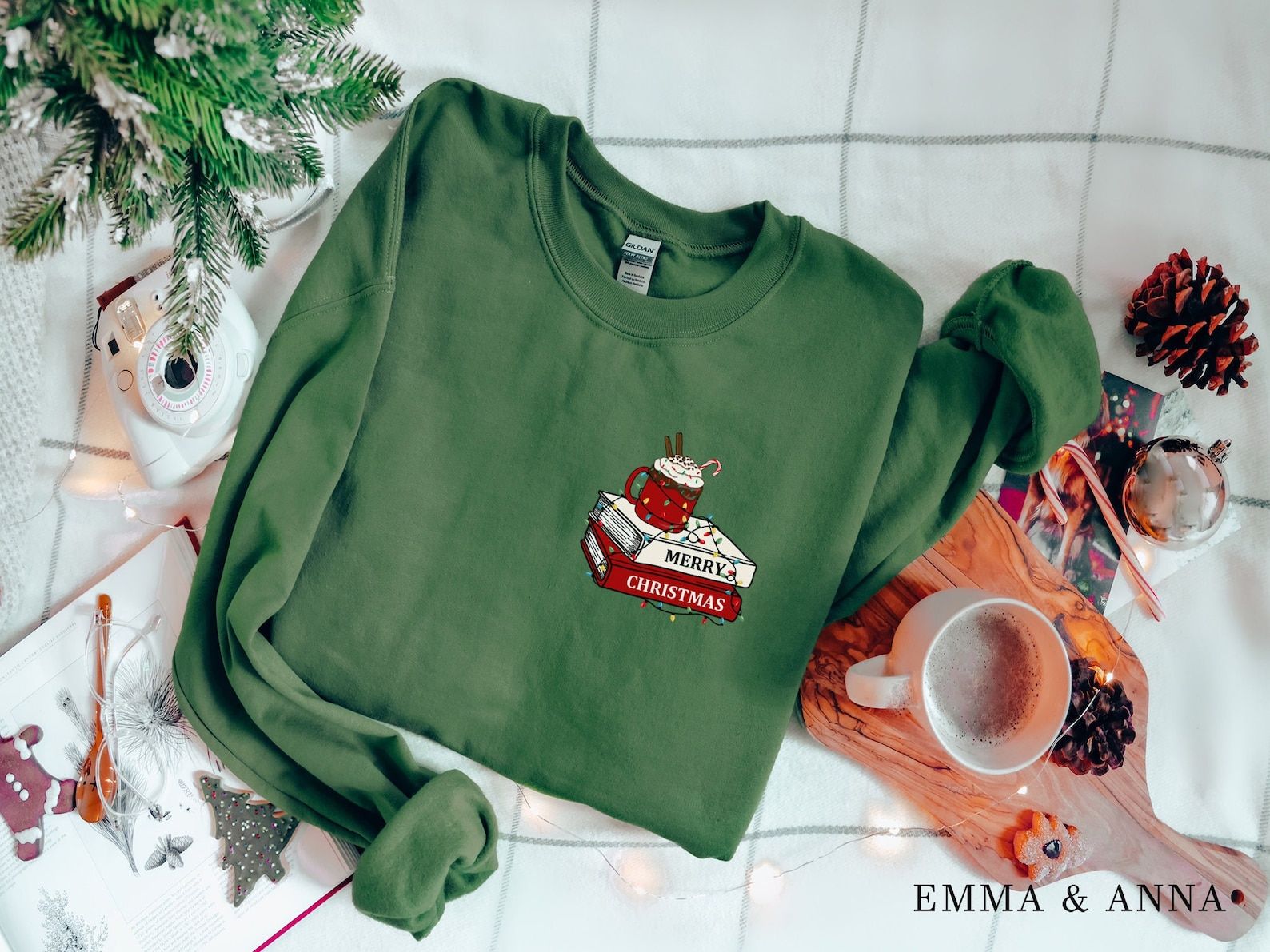 a green sweatshirt with a stack of books and cup of cocoa tangled in lights at the left breast