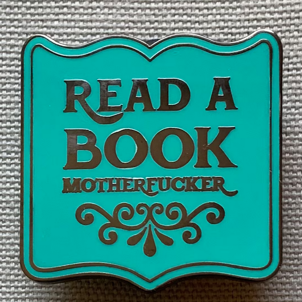 an enamel pin shaped like an opened book with black text that says "read a book motherfucker"