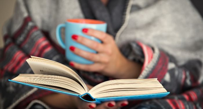 a beige-skinned person with red finger nail polish is holding a mug in one hand and a book in the other; there is a blanket wrapped around them
