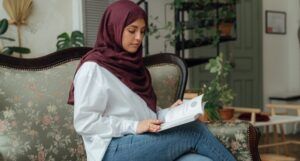 a lightly-tan skinned woman wearing a hijab and reading a book on a couch