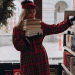 a white woman in red plaid with a stack of books, reaching for another book on the shelf in a bookstore.