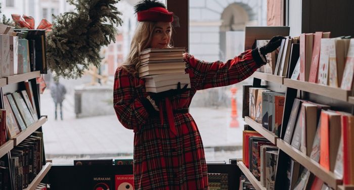 a white woman in red plaid with a stack of books, reaching for another book on the shelf in a bookstore.