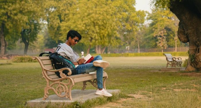 a young brown-skinned South Asian man reading a book on a bench