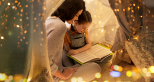 a photo of a mother and daughter reading a book inside of blanket tent strung with lights