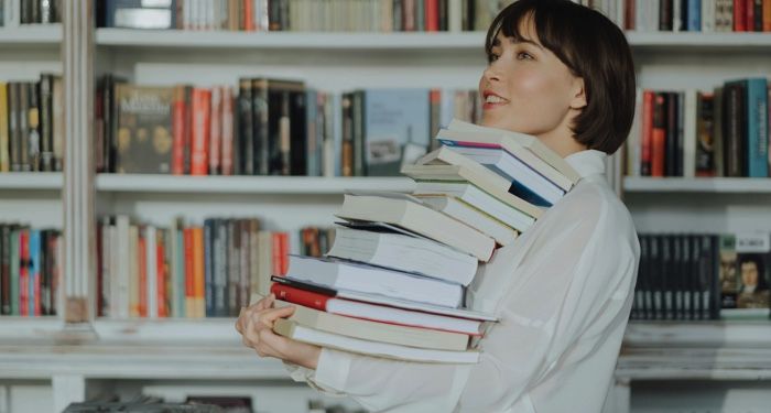 fair-skinned Asian woman smiling and holding a huge stack of books