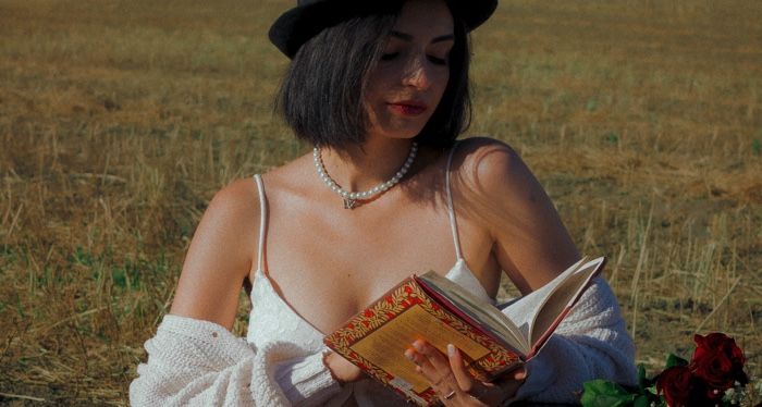 lightly tanned-skinned woman with a black hat on reading in a field next to a bouquet of roses