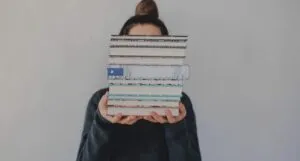 a fair-skinned person with their hair in a bun holding a stack of books that is obscuring their face