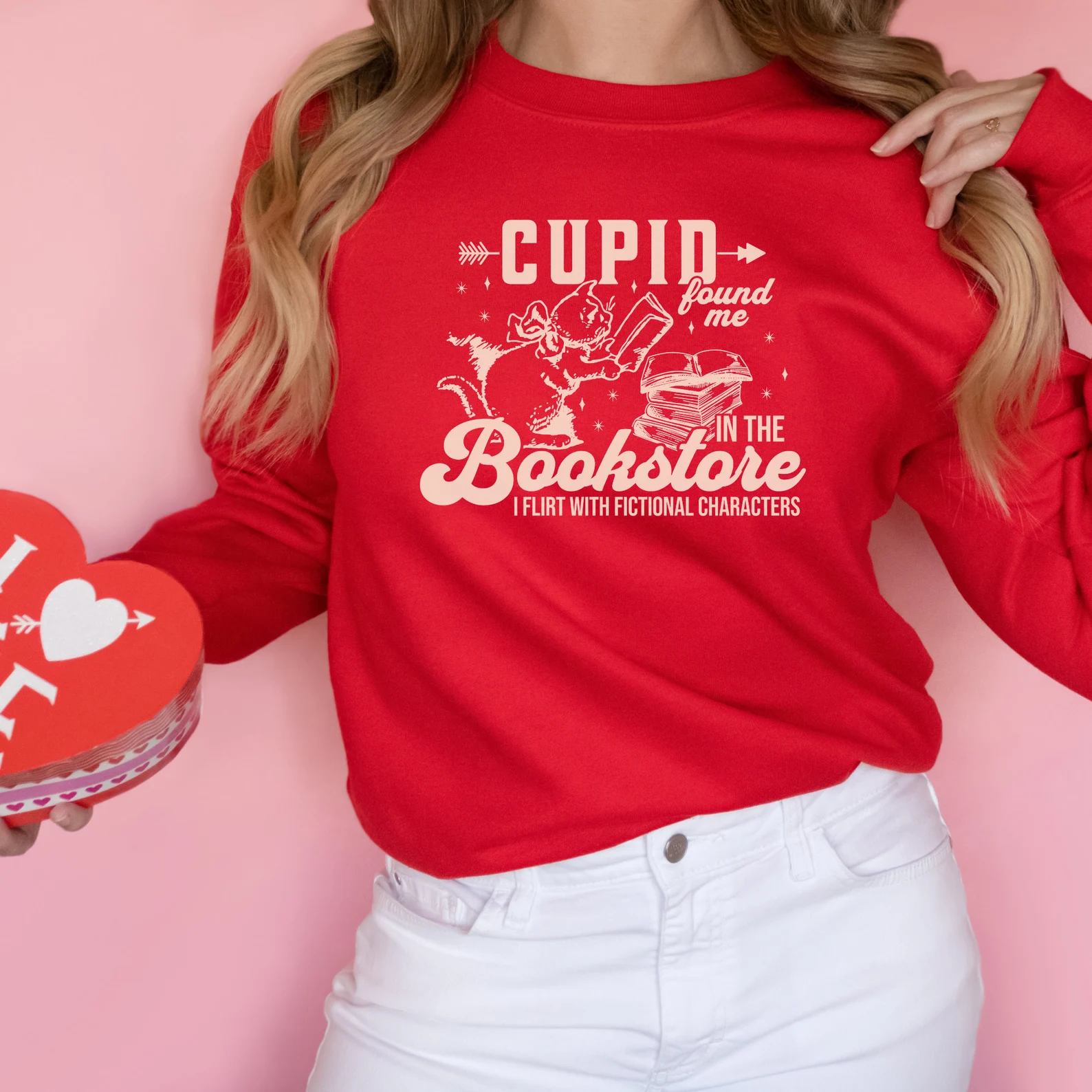 Image of a white person wearing a red  sweatshirt that says "cupid found me in the bookstore. I flirt with fictional characters."