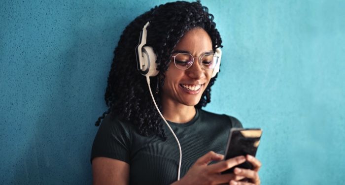 light brown-skinned Black woman with thick curly hair is wearing headphones, looking at her phone, and smiling