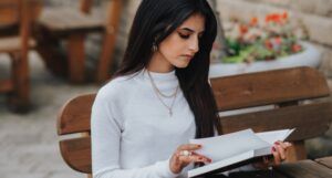 medium fair-skinned woman with long, dark hair reading a book outside at a table
