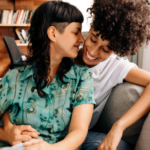 a photo of a queer couple cuddling and smiling in front of a bookcase