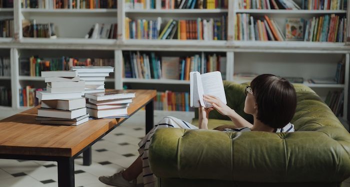a photo of a woman reading on a couch with bookshelves in the background and stacks on books on a table
