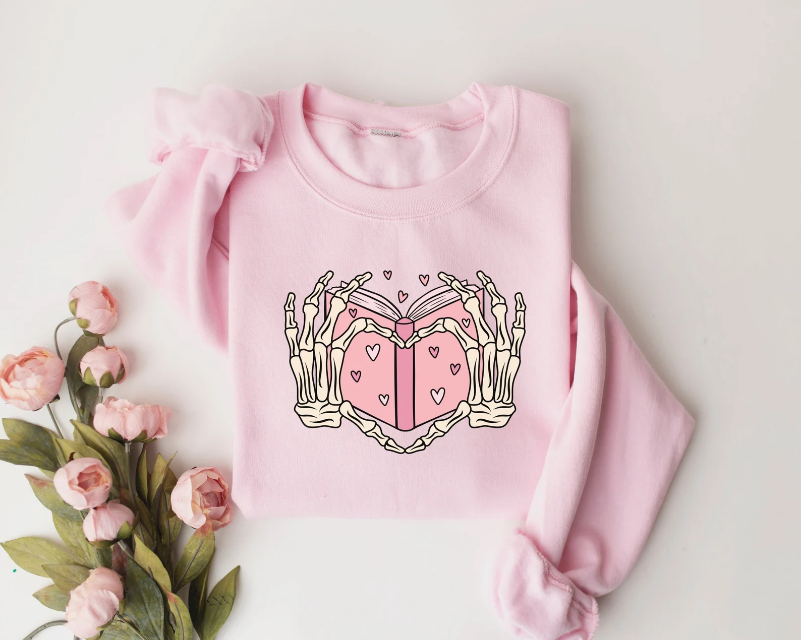 Image of a pink sweatshirt with a heart shape made by two skeleton hands. Between the hands is a book. 