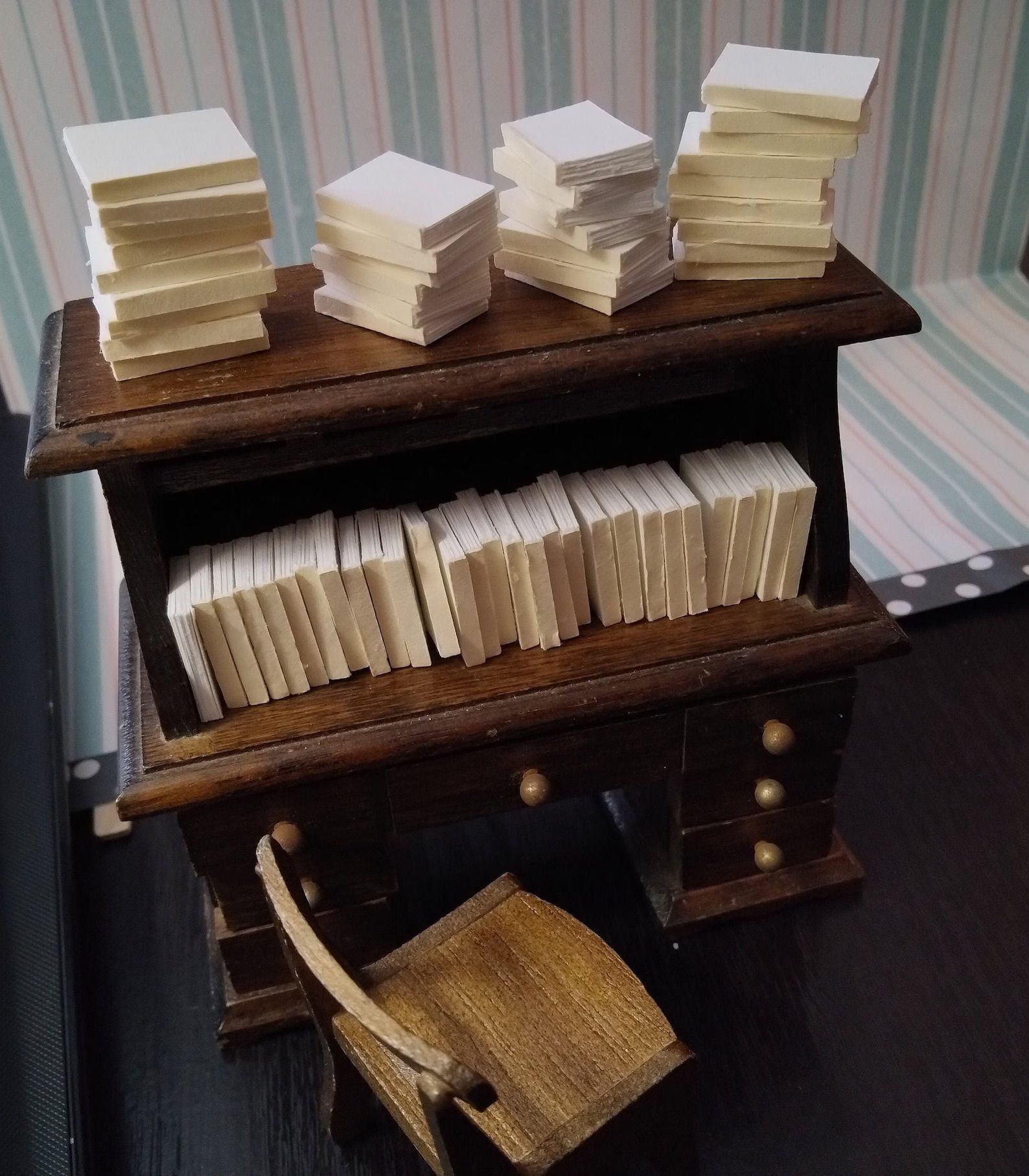 Mini books are in stacks on a mini wooden desk with a mini chair in front of it.