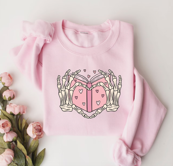 Pink long-sleeve shirt with a design of a pink book with differently colored pink hearts and skeleton hands making a heart shape. 