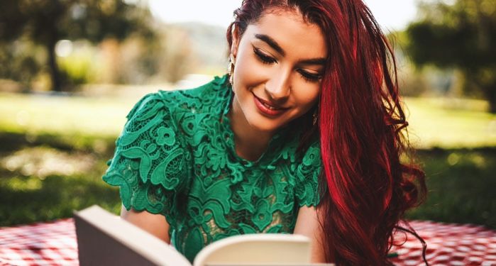 a lightly-tanned skinned woman with long, burgundy hair is lying n a picnic blanket reading a book