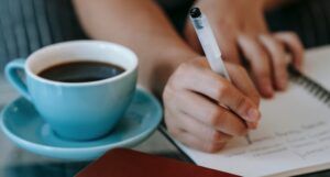 a tan-skinned hand writing in a notebook with a cup of coffee next to them