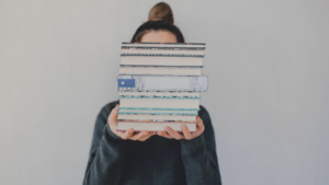 a fair-skinned femme-looking person holding a stack of books that is obscuring their face