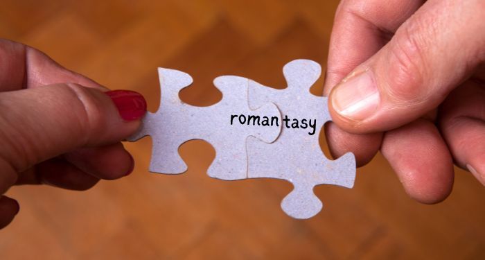 hands holding two connected puzzle pieces. one contains the letters r-o-m-a-n and the other t-a-s-y, combining to spell romantasy