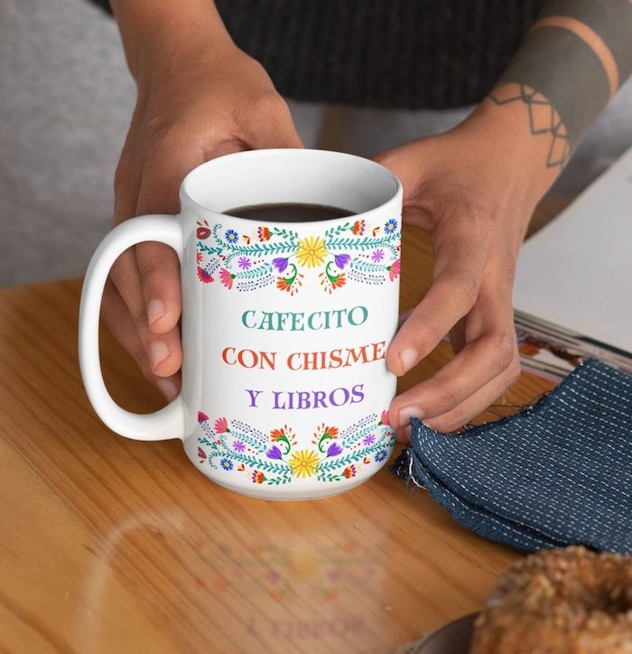 a mug with a painted floral design along the top and bottom. Between the florals are the words "cafecito con chisme y libros"