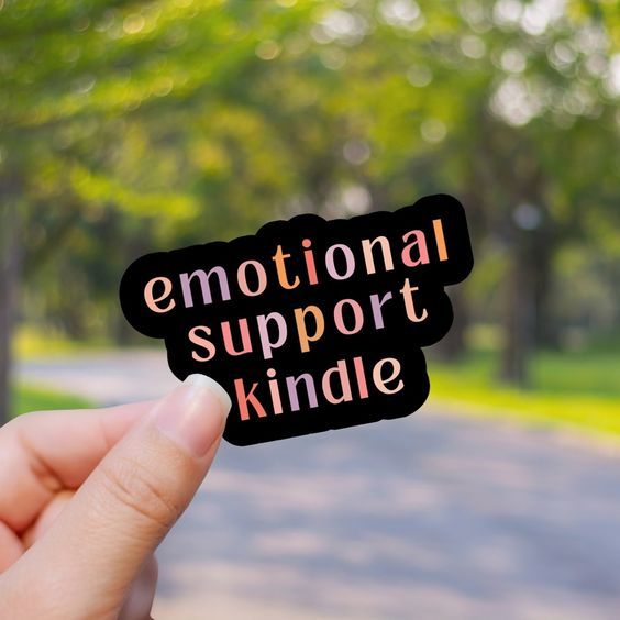 a hand holding a sticker that has pastel letters spelling out emotional support kindle on a black background 