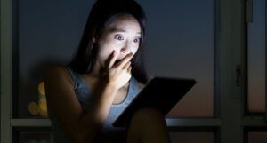 woman holding her hand up to her face in shock while reading from a tablet in the dark