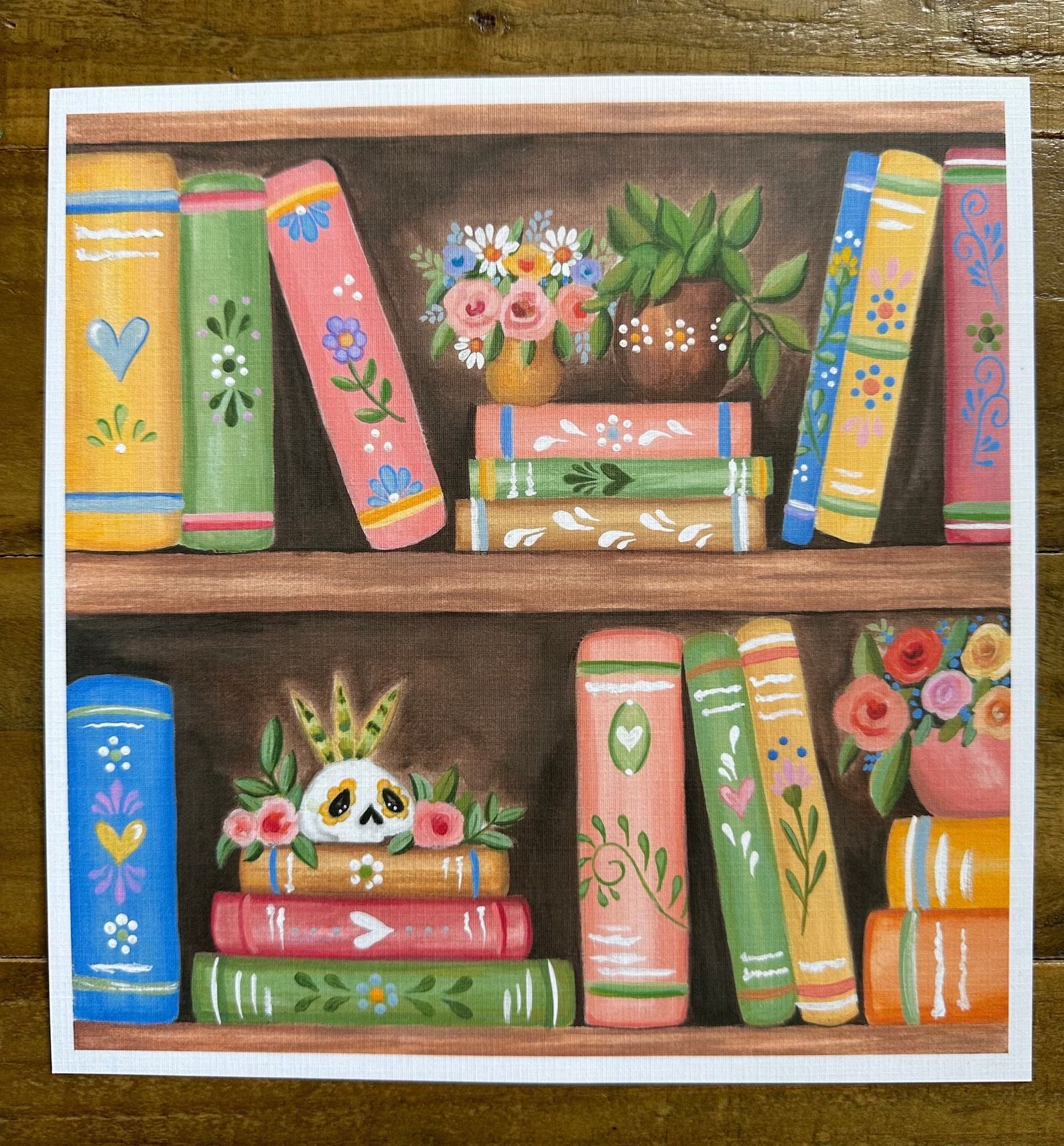 a print showing a bookshelf with several colorful books, succulents, and a painted skull 