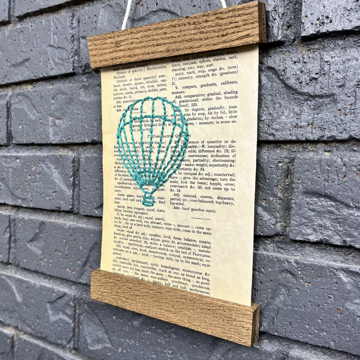 Image of an old book page embroidered with a teal hot air balloon hanging from a brick wall with a wooden frame