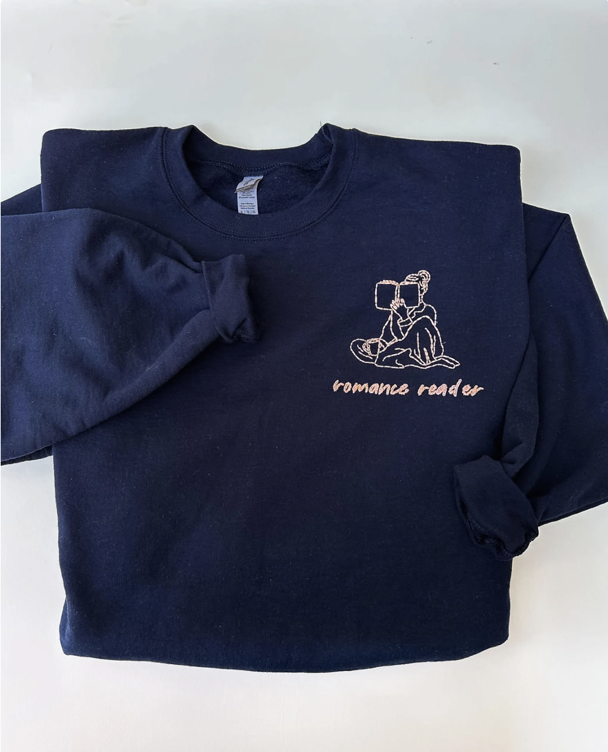 Image of a navy sweatshirt with embroidery on the left chest of a woman reading a book and the text "romance reader"