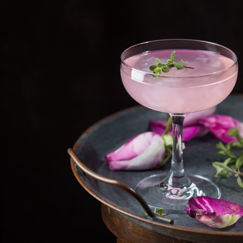 a lilac-colored cocktail in a glass, surrounded by roses