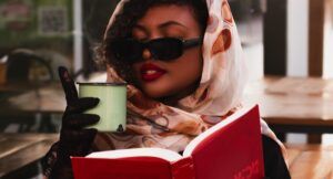 a fabulous brown-skinned Black woman wearing a head scarf over her curly hair, holindg a book, and a hot beverage