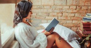 a tanned skin woman sitting upright in a bed and reading a book