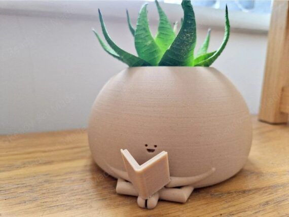 a beige round planter with a tiny face a body holding an open book with a succulent peeking out of the top