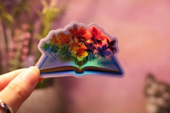 small holographic sticker in the shape of an open book with flowers growing out the pages 