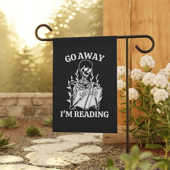 small black garden flag with the words "go away I'm reading" and a skeleton reading surrounded by flowers 