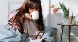 a medium-skinned Asian little woman drinking coffee and reading a book in bed