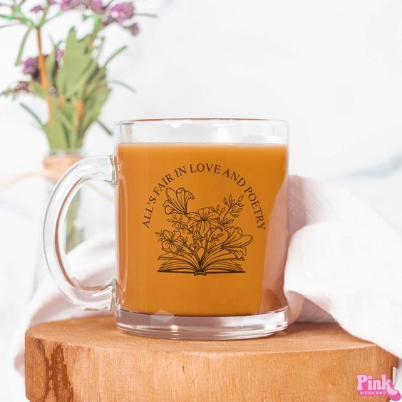 a clear glass mug with black lettering and a black line drawing of flowers blooming out of an open book. The lettering reads "All's Fair in Love and Poetry"
