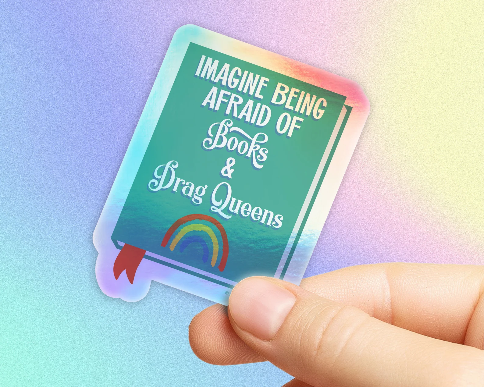 A holographic sticker that is in the shape of a book. On the cover it says "imagine being afraid of books and drag queens."
