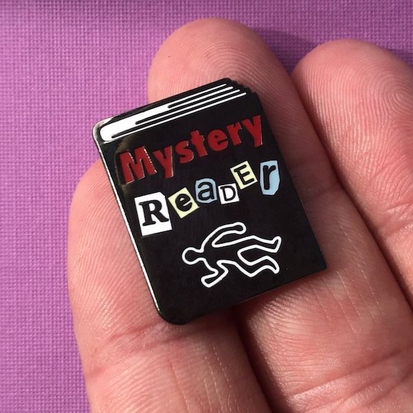 an enamel pin in the shake of a black book. the front cover of the book contains. text that reads "mystery reader" hovering over the chalk outline of a body