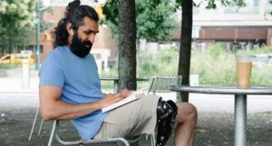 olive-skinned man with long, dark hair and a prosthetic leg is reading while sitting at a table outside