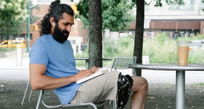 olive-skinned man with long, dark hair and a prosthetic leg is reading while sitting at a table outside