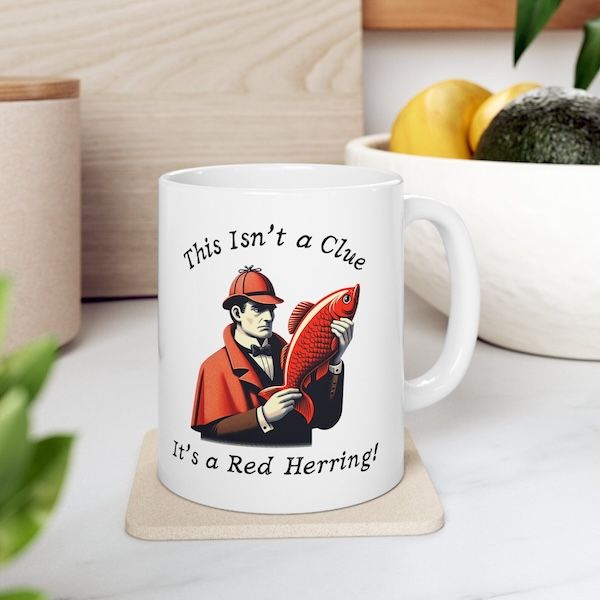white coffee mug with a graphic of a detective holding a red fish. Text surrounding the image reads "This Isn't a Clue, It's a Red Herring!"I