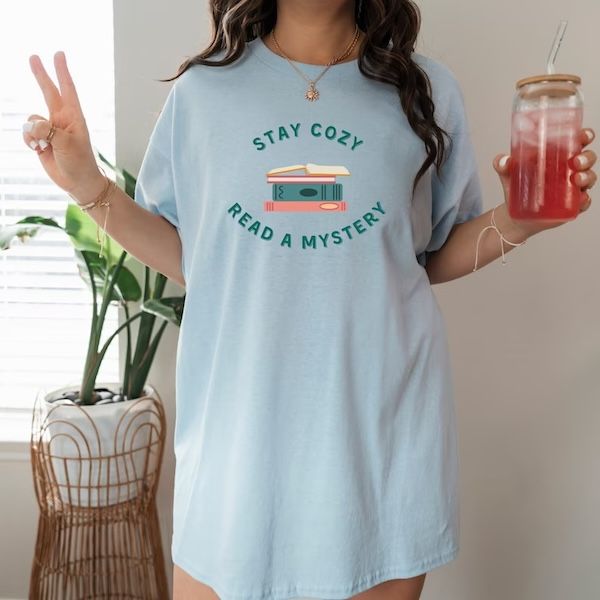 woman wearing a light blue tshirt. The front of the shirt contains a graphic of a small stack of books and text that reads "Stay cozy, read a mystery"