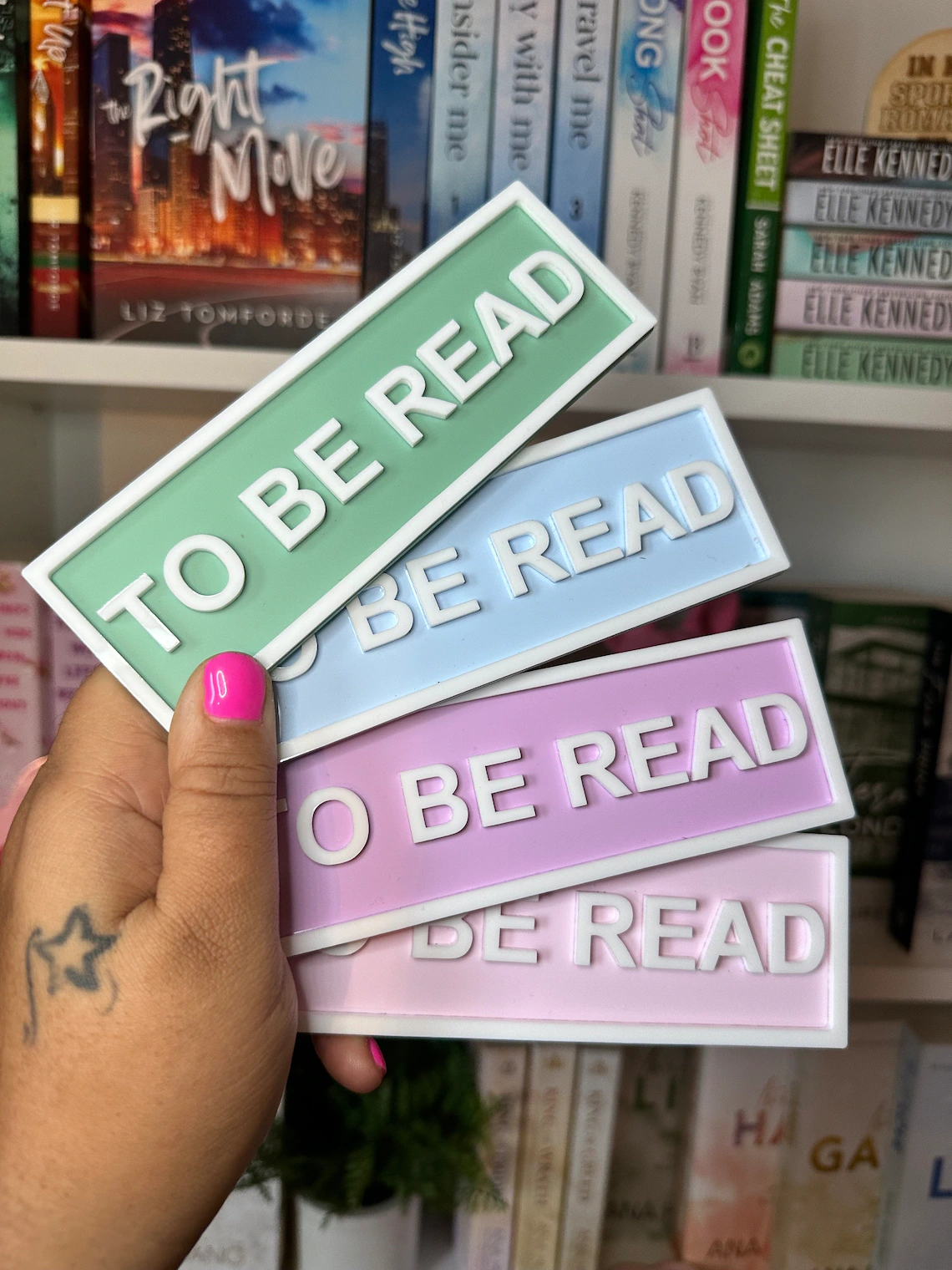Three To Be Read signs in pastel colors being held by a hand