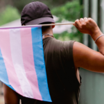 a person facing away from the camera holding a trans pride flag behind them