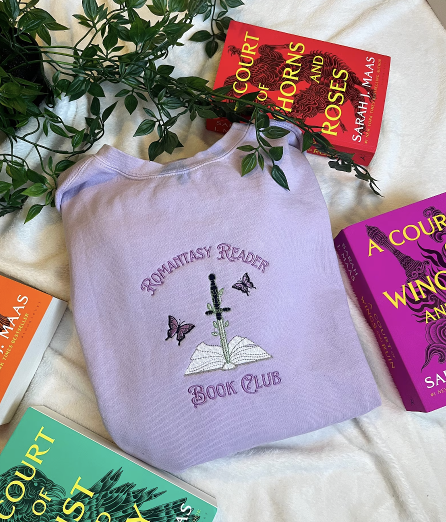 Image of a light purple sweatshirt with a book, sword, and dragons and the saying "romantasy reader book club" embroidered on it, folded on a white background surrounded by fantasy romance books
